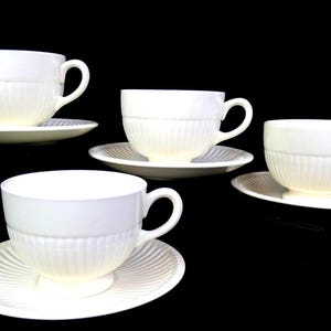 Op de afbeelding: Vier witte theekopjes en schotels tegen een zwarte achtergrond. De kopjes hebben een geribbeld ontwerp en gebogen handvatten. De schotels hebben ook een geribbeld patroon. Een klassieke theeset.
