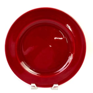 Könnte beinhalten: Ein runder, glänzend roter Teller mit breitem Rand. Der Teller wird auf einem transparenten Kunststoffständer vor weißem Hintergrund präsentiert. Die Oberfläche des Tellers reflektiert das Licht.
