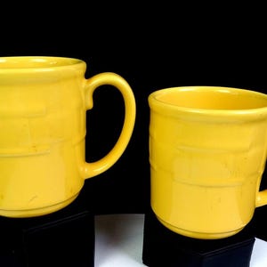May include: Two bright yellow ceramic mugs with a smooth, glossy finish. Each mug features a distinct horizontal embossed band pattern around the center of the body, a sturdy curved handle on the side, and a wide, circular rim at the top.