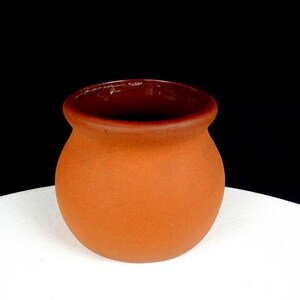Puede incluir: Una pequeña maceta de terracota redonda con una boca ancha y abierta y un acabado exterior liso y mate. El recipiente presenta un borde distintivo ligeramente acampanado y un esmalte interior de color marrón oscuro que contrasta con la arcilla.