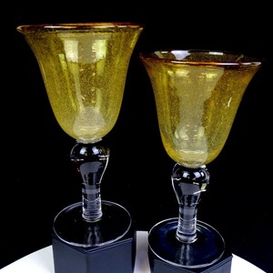 May include: Two matching goblets featuring golden-yellow bowls with a subtle speckled texture and a dark amber rim. Each glass has a clear, multi-faceted stem and a wide, circular base, providing a stable foundation for the flared drinking vessel.