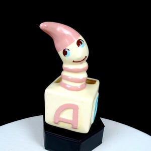 May include: A ceramic figurine featuring a pink and white worm emerging from a cream-colored alphabet block. The block displays a large pink letter A on the front. The worm has a curved pink head, blue eyes, and a segmented body resting on the block.