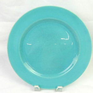 May include: A round, solid turquoise ceramic plate with a smooth, glossy finish and a wide, flat rim. The plate features a simple, circular design with a slightly recessed center, showcasing a vibrant, uniform color across the entire surface.