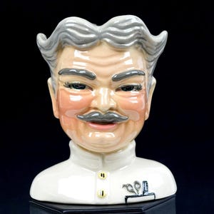 Może przedstawiać: Ceramiczny dzbanek w kształcie postaci z siwymi włosami, siwymi wąsami i w białym mundurze. Figurka posiada wysoki kołnierz, dwa żółte guziki oraz małą grafikę przedstawiającą nożyczki i grzebień w okolicy klatki piersiowej.