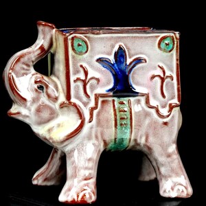 May include: A ceramic planter shaped like an elephant with a square body. The piece features a glossy finish in shades of light brown, cream, and white, with a prominent blue fleur-de-lis emblem and small green accents on the sides of the body.