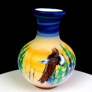 May include: A round ceramic vase featuring a hand-painted scene of a bald eagle in flight against a large yellow sun. The background includes green pine trees and a gradient sky that transitions from a soft yellow to a deep blue at the neck.
