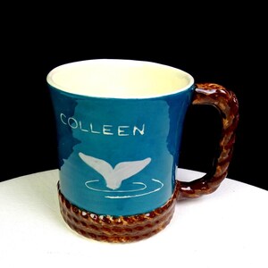Może przedstawiać: Ceramiczny kubek z niebieskim nadrukiem z napisem "COLLEEN" i białym wzorem ogona wieloryba. Uchwyt jest brązowy i przypomina linę, a podstawa ma pasujący brązowy wzór liny. Wnętrze jest białe.