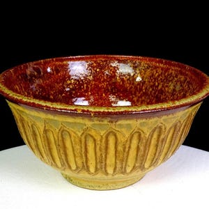 May include: A ceramic bowl featuring a deep, glossy reddish-brown interior with a speckled texture. The exterior is a muted mustard yellow with a repeating vertical fluted or carved pattern around the sides, ending in a smooth, circular base.