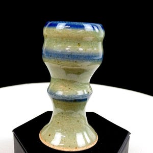 May include: A ceramic goblet with a glossy finish, featuring a light green base color with horizontal bands of deep blue glaze. The surface has a visible crackle texture throughout, and the vessel has a distinct hourglass shape with a flared rim.