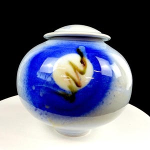 May include: A ceramic urn with a lid, featuring a vibrant blue and white color scheme. The urn has a spherical shape with a circular lid. The design includes a central cream-colored motif with brown and black accents.