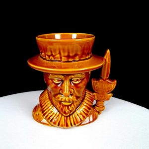 May include: A glossy, amber-colored ceramic mug shaped like a bust of a person wearing a wide-brimmed hat and a ruffled collar. The handle of the mug is designed to look like a vertical sword or dagger, extending upward from the side of the base.