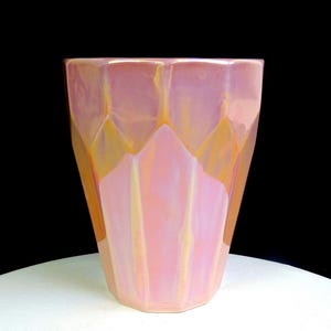 May include: A faceted ceramic vase with a glossy, iridescent finish that shifts between soft pink, peach, and golden yellow tones. The vessel features a geometric, multi-sided structure with vertical panels that taper toward the base.