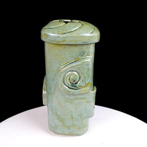 May include: A ceramic vase with a sage green glaze and a glossy finish. The vessel features a rectangular body with rounded corners, a lid with a swirling embossed pattern, and a prominent spiral design on the front face. The surface shows tonal variations.