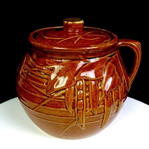 Puede incluir: Una olla de cerámica marrón con tapa y asa. La olla tiene una superficie texturizada con un diseño de hojas. La tapa tiene un pequeño pomo. La olla está sobre una superficie blanca con un fondo negro.