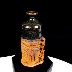 May include: A ceramic bottle with a glossy black upper section and a textured, matte terracotta-coloured lower section. The base features intricate, hand-carved geometric patterns and a vertical, rugged, decorative ridge along one side.