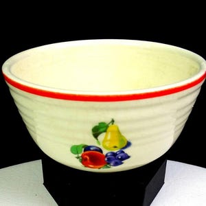Può includere: Ciotola in ceramica color crema con bordo rosso e disegno di frutta. La ciotola presenta una pera, una mela e dell'uva. La ciotola ha una trama a coste ed è posta su uno sfondo nero.
