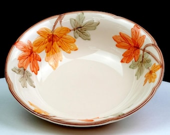 May include: A round ceramic bowl with a cream-coloured base and a scalloped rim. The interior features a painted pattern of orange and muted green maple leaves with brown stems. A thin brown line accents the outer edge of the bowl's rim.