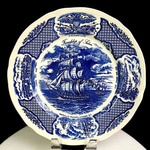 May include: A cream-colored ceramic plate featuring a blue transferware design. The center depicts the ship Friendship of Salem. The border includes an eagle, the text E Pluribus Unum, and scenes labeled Canton Port 1830, New York Harbor 1830, and U.S. Delaware Capturing Le Croyable.