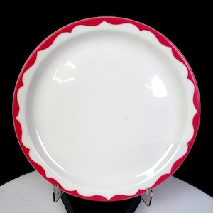 May include: A white ceramic plate with a scalloped red rim. The plate is round and has a glossy finish. The red rim has a repeating wave pattern. The plate is displayed on a clear stand against a black background.