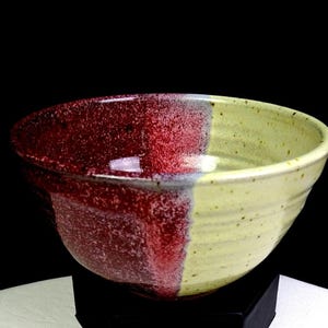 Puede incluir: Un cuenco de cerámica hecho a mano con un diseño de dos tonos. Una mitad es de color burdeos moteado, la otra es de color amarillo pálido moteado. El cuenco tiene forma redondeada y probablemente se utiliza para servir alimentos.