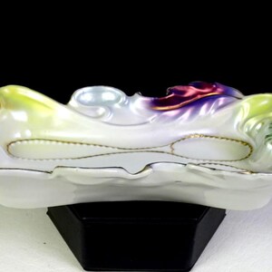 May include: A white porcelain ashtray with a scalloped edge and a decorative, iridescent floral design in shades of yellow, green, purple, and gold. The ashtray has a small indentation for resting a cigarette.