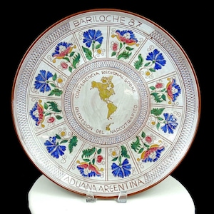 Può includere: Un piatto in ceramica bianca con un bordo marrone e un design decorativo di fiori in tonalità di rosso, blu e verde. Il centro del piatto presenta una mappa delle Americhe con il testo "CONFERENCIA REGIONAL SOBRE REPRESION DEL NARCOTRAFICO" e "BARILOCHE 87" e "ADUANA ARGENTINA".