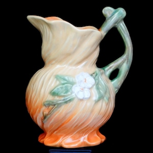 May include: A ceramic pitcher featuring a swirling, ribbed texture with a gradient color scheme transitioning from a pale cream at the top to a vibrant orange at the base. A green handle is attached to the side, and a white floral relief.