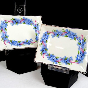 May include: Two rectangular porcelain dishes with scalloped edges and a gold-tone trim. The center of each dish is white, surrounded by a decorative border of blue forget-me-not flowers, green leaves, and flowing pink ribbons on a cream background.