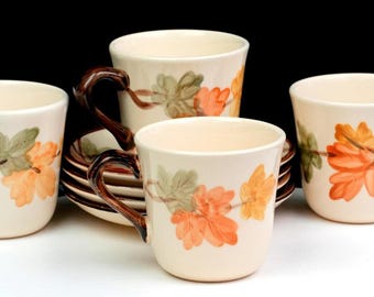May include: A set of four cream-coloured ceramic mugs and matching saucers featuring a painted design of orange and green leaves on brown stems. Each mug has a dark brown handle and a smooth, rounded body, arranged in a cluster against a black background.