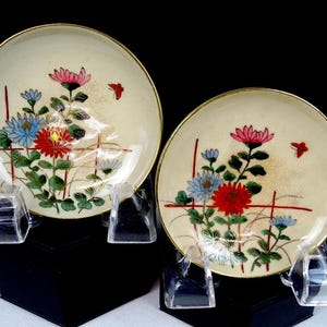 Peut inclure: Deux assiettes rondes en céramique blanc cassé avec des bords dorés, chacune présentant un motif floral peint à la main de chrysanthèmes rouges, bleus et roses, des feuilles vertes et un petit papillon rouge sur une grille de lignes rouges.