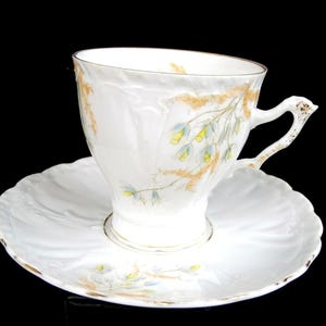 以下が含まれることがあります： 白のティーカップとソーサーのセット。黄色と青の繊細な花柄のデザイン。ティーカップはカーブしたハンドルと金のトリムが付いています。ソーサーはスカラップエッジで、ティーカップのデザインと一致します。黒い背景。