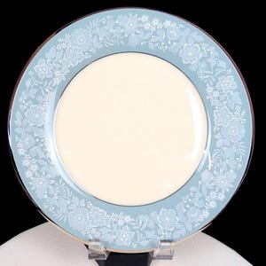 May include: A round ceramic dinner plate featuring a solid cream-colored center and a wide light blue border. The border is decorated with a repeating white floral pattern consisting of various blossoms, leaves, and small sprigs around the rim.