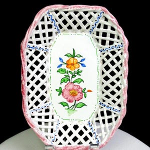 May include: An oval ceramic basket with a white lattice-patterned wall and a pink scalloped rim. The center base features a hand-painted floral design with a large pink flower, a smaller orange flower, and green leaves, accented by blue wavy lines.