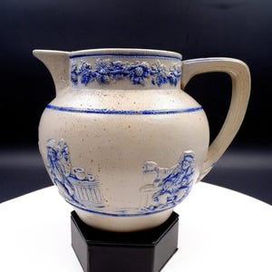 Może przedstawiać: Kamionkowy dzban z pękatym korpusem i uchwytem. Dzban jest beżowy z niebieskimi reliefami przedstawiającymi postacie i wzory kwiatowe. Niebieski pas otacza górę i podstawę. Dzban jest eksponowany na czarnym stojaku.