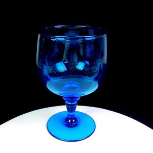 May include: A vibrant cobalt blue glass goblet featuring a rounded bowl, a short stem with a central decorative node, and a wide circular base. The glass is translucent, allowing light to pass through the deep blue material, creating highlights.