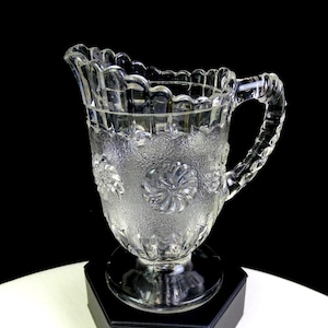 Könnte beinhalten: Klarer Glaskrug mit strukturierter Oberfläche und floralen Designs. Der Krug hat einen gewellten Rand und einen dekorativen Griff. Der Krug steht auf einem schwarzen Sockel.