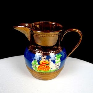 May include: A small, antique ceramic pitcher with a brown metallic glaze on the top and handle. The base is blue with a painted scene of figures and foliage. The pitcher has a spout and a curved handle.