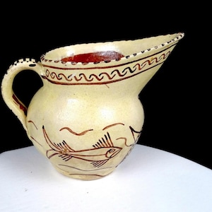 以下が含まれることがあります： クリーム色の陶器製ピッチャーで、胴体には手描きの茶色の魚のモチーフ、縁には波線の模様が施されています。ピッチャーには湾曲したハンドルと広い注ぎ口があり、内側は濃い赤茶色で仕上げられています。