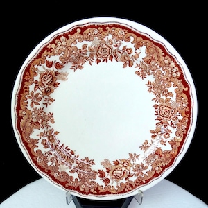 May include: A white ceramic plate with a detailed floral pattern in shades of brown and red. The plate has a scalloped edge and a decorative border, showcasing a classic design. This vintage-style plate is suitable for serving food.