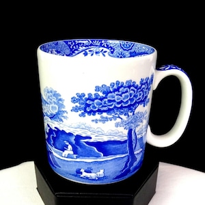 Puede incluir: Taza de cerámica blanca con un diseño de paisaje azul. La taza presenta una ilustración detallada en azul y blanco de un paisaje escénico con árboles, figuras y un borde decorativo. El asa también está decorada con detalles azules.