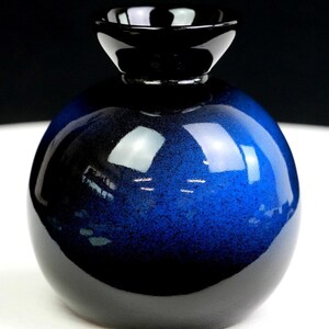 Puede incluir: Un jarrón esférico de vidrio con un degradado azul profundo que transiciona hacia una base negra sólida y un borde negro ancho y acampanado. La superficie tiene un acabado brillante y reflectante con una textura moteada en el azul.