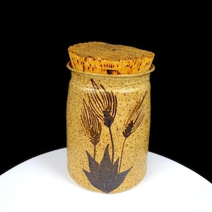 Op de afbeelding: Een cilindrische keramische pot met een gespikkelde beige afwerking en een donkerbruin handgeschilderd ontwerp van tarwearen. De houder heeft een brede opening die is afgesloten met een dikke, natuurkleurige kurken stop.