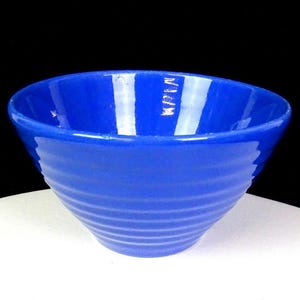 Puede incluir: Un cuenco de cerámica azul vibrante con acabado brillante. El cuenco tiene forma redondeada con un borde ligeramente ensanchado y detalles horizontales acanalados en el exterior. El cuenco está sobre un fondo negro.