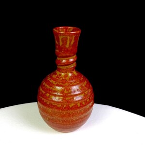 Può includere: Un vaso in ceramica con corpo sferico e collo stretto a spirale. La superficie presenta una smaltatura maculata nei toni dell'arancione vibrante e del rosso intenso, con striature orizzontali sulla base rotonda e un bordo superiore liscio.