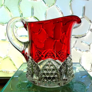 May include: A glass pitcher featuring a vibrant red upper section and a clear, textured base with a geometric diamond pattern. The pitcher includes a curved handle on the side and a wide pouring spout, showcasing a distinct contrast between the smooth red glass and the intricate, faceted clear glass bottom.