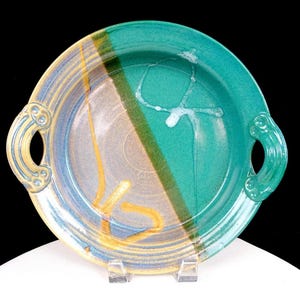 May include: A round ceramic serving dish with two side handles, featuring a split color design of teal and a muted tan-blue blend. The surface displays a glossy finish with abstract yellow linear accents and a white drip pattern on the teal side.