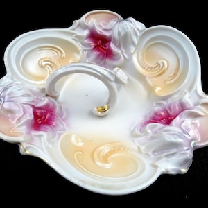 May include: A white porcelain serving dish with a handle. The dish features embossed floral designs with pink and yellow accents. The handle has a gold-coloured detail.