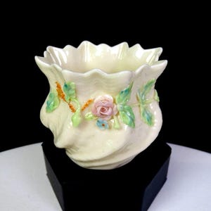 May include: A cream-colored ceramic vessel with a scalloped rim and a pearlescent finish. The exterior is decorated with raised, hand-painted floral details, including a central pink rose, green leaves, and small blue flowers on a vine.