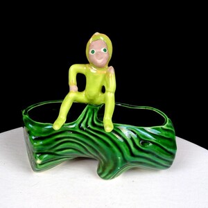 Può includere: Un'antica fioriera in ceramica con una figura seduta in tuta verde lime. La figura ha un volto sorridente ed è appollaiata su una fioriera verde con un design strutturato simile al legno. La fioriera ha due aperture.