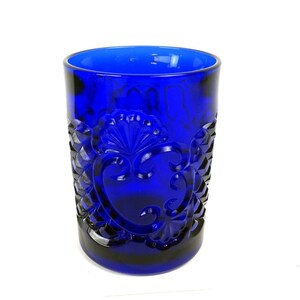 May include: A vibrant cobalt blue drinking glass with an embossed design. The glass has a detailed pattern of stylized leaves and a central heart-shaped motif. The glass is cylindrical and has a thick base.